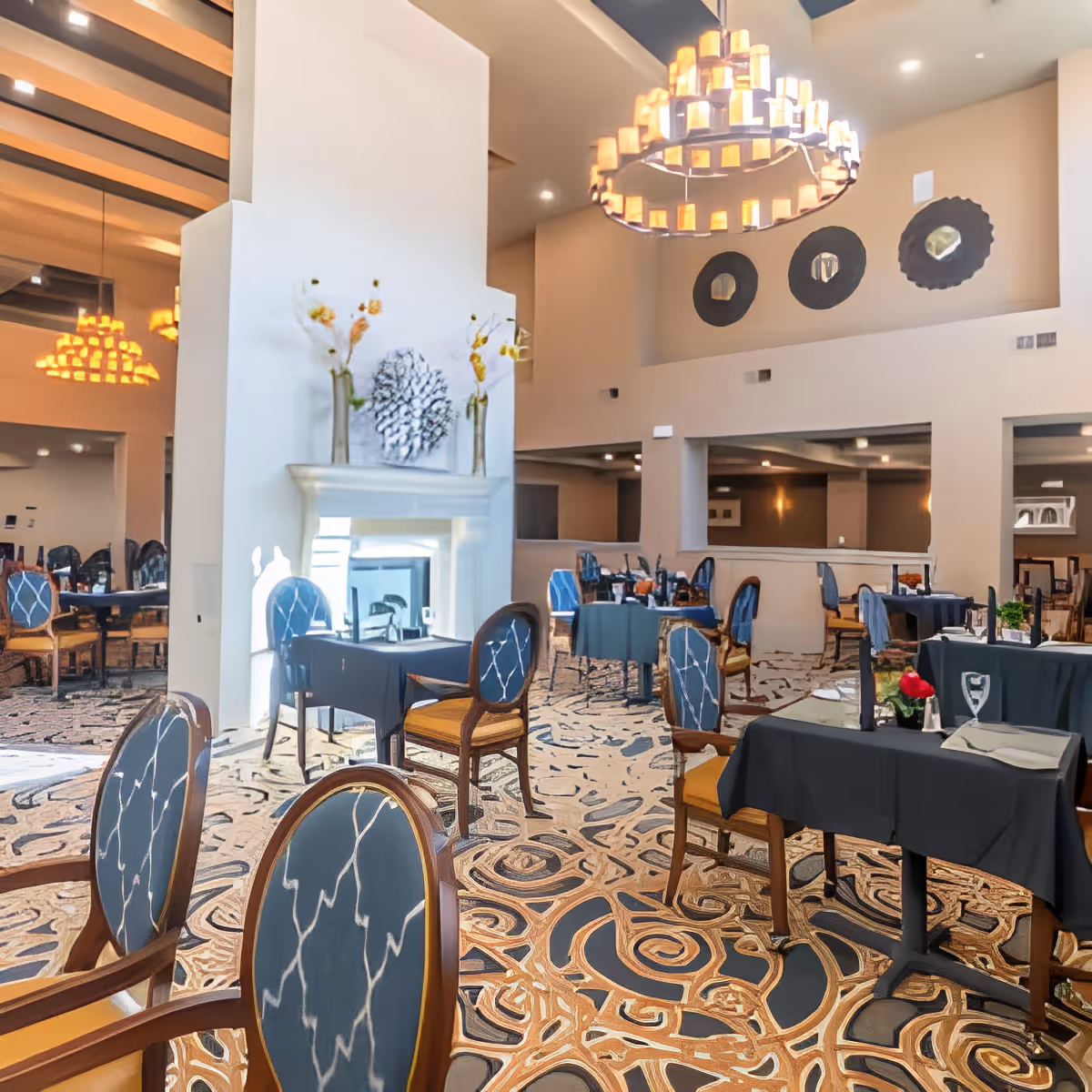 A spacious dining room with multiple tables covered in dark tablecloths and set with plates and glasses. The room features elegant wooden chairs with blue patterned upholstery, a large decorative chandelier, a fireplace with vases and artwork above it, and a patterned carpet. The walls are painted in neutral tones with decorative circular wall hangings.