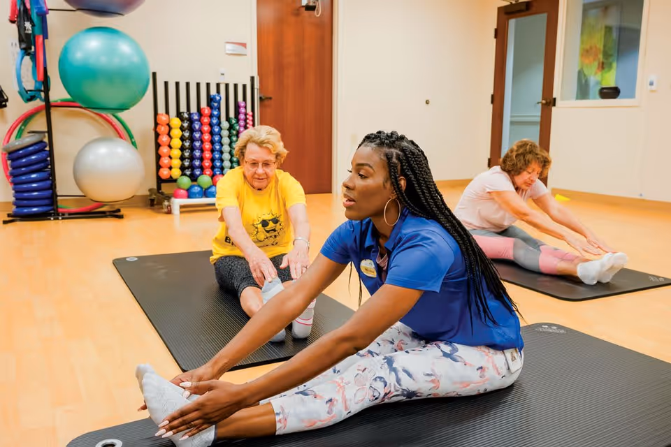A fitness instructor leads two elderly women in a seated forward stretch exercise on black mats in a well-lit exercise room with wooden floors. Various colorful exercise equipment, including stability balls, dumbbells, and hula hoops, are visible in the background.
