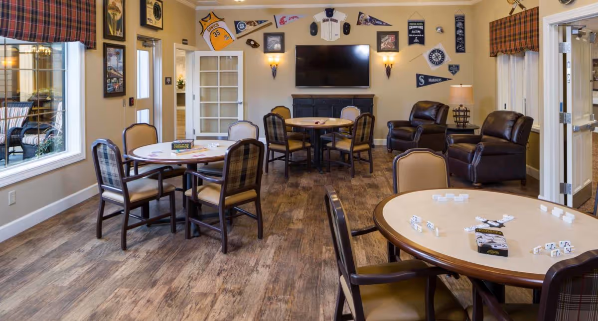 A cozy common area in Cedar Creek Memory Care Community featuring round tables with chairs arranged for games and socializing. The room has wood flooring, a wall-mounted TV above a fireplace, and sports memorabilia decorating the walls. Large windows with plaid valances let in natural light, and there are comfortable leather armchairs near a lamp in the corner.
