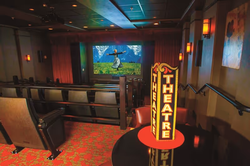 Interior view of a small theater room with rows of cushioned seats facing a large screen showing a scene from The Sound of Music. A decorative illuminated sign reading 'THEATRE' is placed on a round table near the entrance. The room has warm lighting and red patterned carpet.