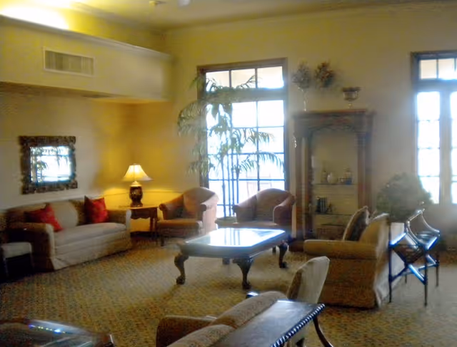 A cozy living room with beige walls and carpet, featuring a sofa with red cushions, two armchairs, a wooden coffee table, a side table with a lamp, a decorative mirror on the wall, a tall plant near large windows, and a wooden shelving unit with decorative items.