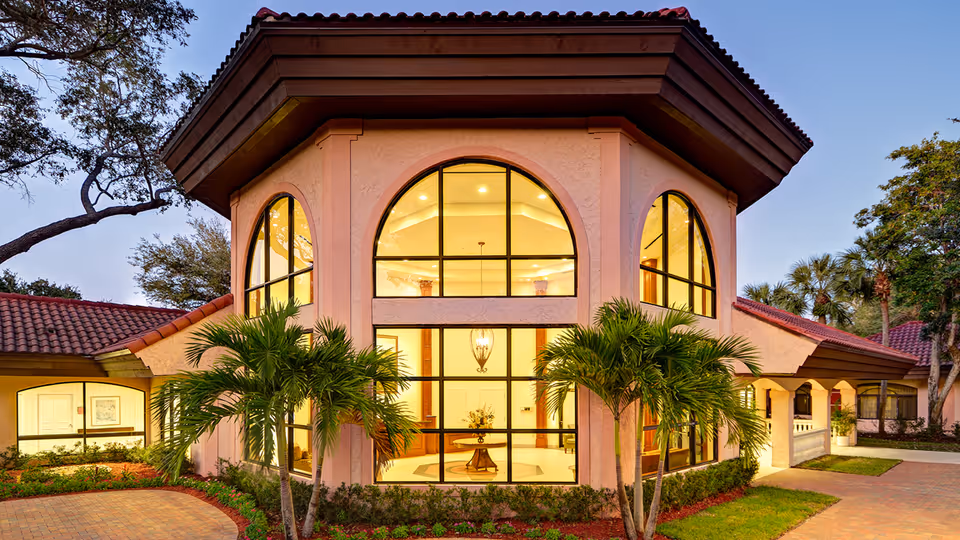 Exterior view of a building at Regents Park Boca Raton with large arched windows, a tiled roof, palm trees, and a paved driveway during dusk.