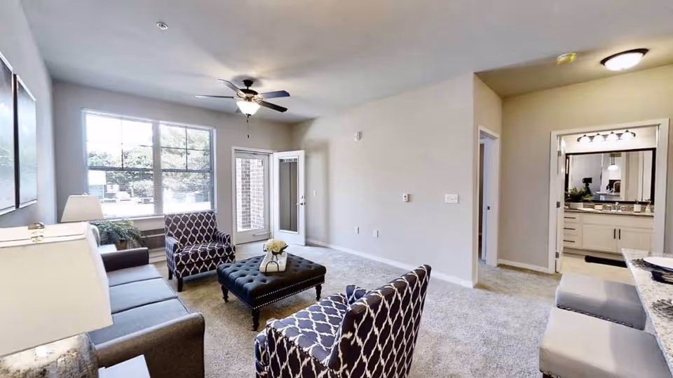 A bright living room with a gray sofa, two patterned armchairs, and a tufted ottoman in the center. There is a ceiling fan with a light fixture, large windows letting in natural light, and a door leading outside. To the right, there is an open doorway leading to a bathroom with a large mirror and vanity, and a kitchen counter with two stools is partially visible.