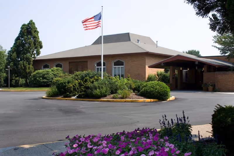 Exterior view of a brick building with a sloped roof, surrounded by greenery and a circular driveway. An American flag is flying on a flagpole in front of the building. There are purple flowers in the foreground.