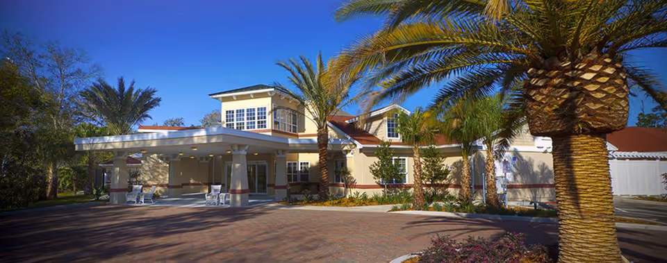 Exterior view of Grand Palms Assisted Living & Memory Care facility with a covered entrance, palm trees, and clear blue sky.