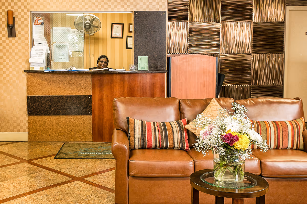 Lobby reception area with a leather sofa and floral centerpiece in front of a wooden reception desk where a person is visible in the window.