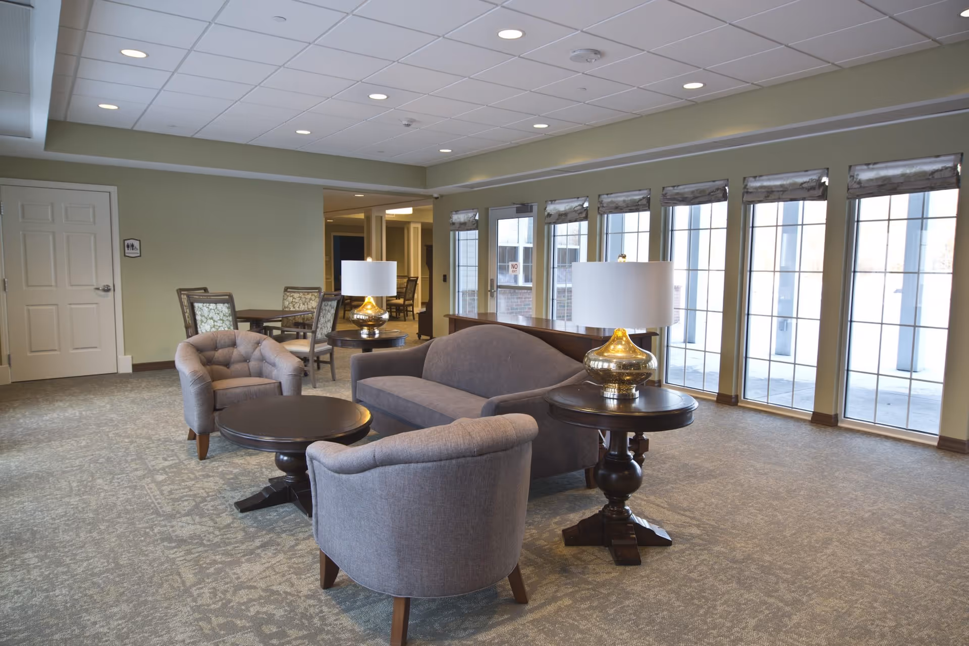 Sunlit communal lounge with gray upholstered chairs and a sofa arranged around round wooden tables and lamps in front of tall windows.