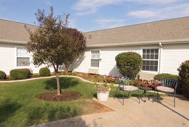 Outdoor patio area at Morada Lake Hefner with a small tree, green grass, flower beds, two metal chairs with cushions, and a small round table on a concrete walkway in front of a white building with windows.
