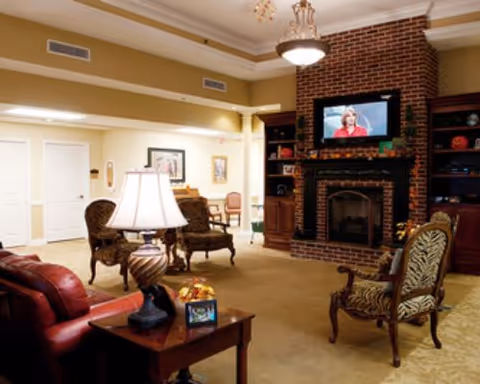 A cozy living room area with a brick fireplace and a mounted TV above it. The room features classic-style armchairs, a wooden side table with a lamp, and warm lighting. There are built-in shelves on either side of the fireplace with decorative items, and the walls are painted in a soft yellow tone.