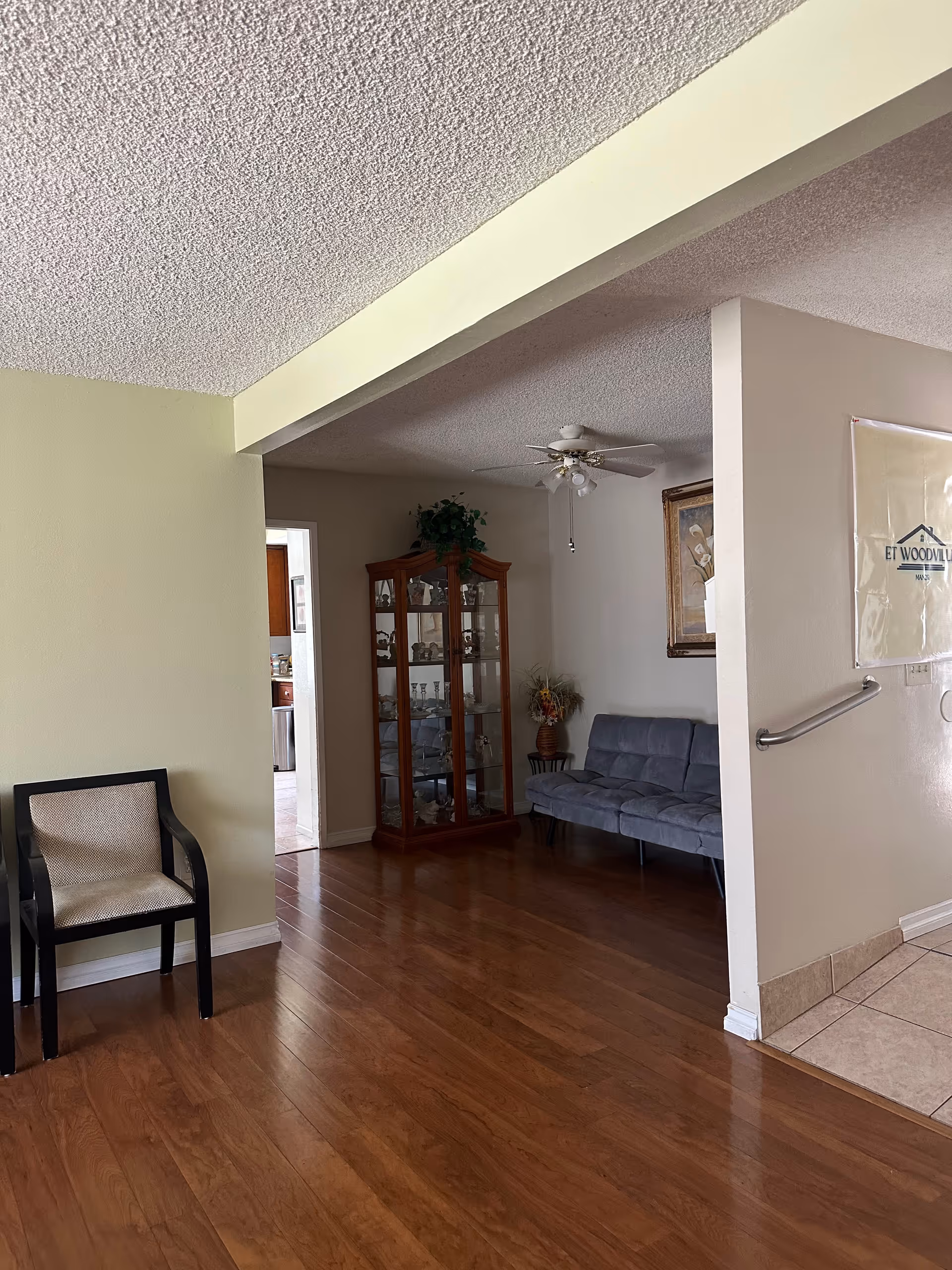 Interior view of a senior living facility showing a wooden floor, a black chair with beige upholstery on the left, a glass display cabinet with decorative items in the center, a gray couch against the wall, a ceiling fan with lights, a framed picture on the wall, and a partial view of a tiled floor area with a handrail and a banner that reads ET WOODVILLE MANOR.