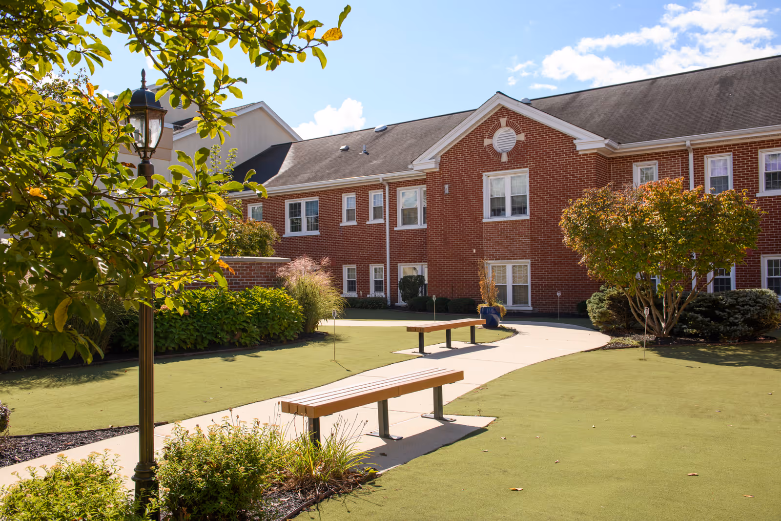 Sunlit courtyard with benches, a paved path, manicured lawn, and a red brick building in the background.