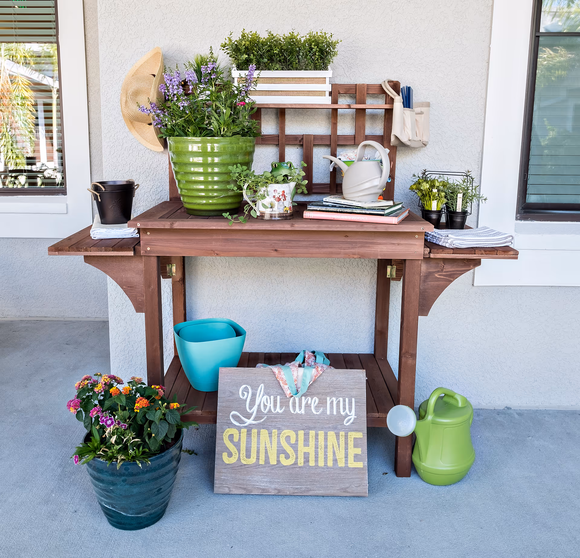 A wooden potting bench against a light gray wall with various gardening items including potted plants, a watering can, gardening books, a hat hanging on the wall, and a decorative sign that reads 'You are my SUNSHINE'.