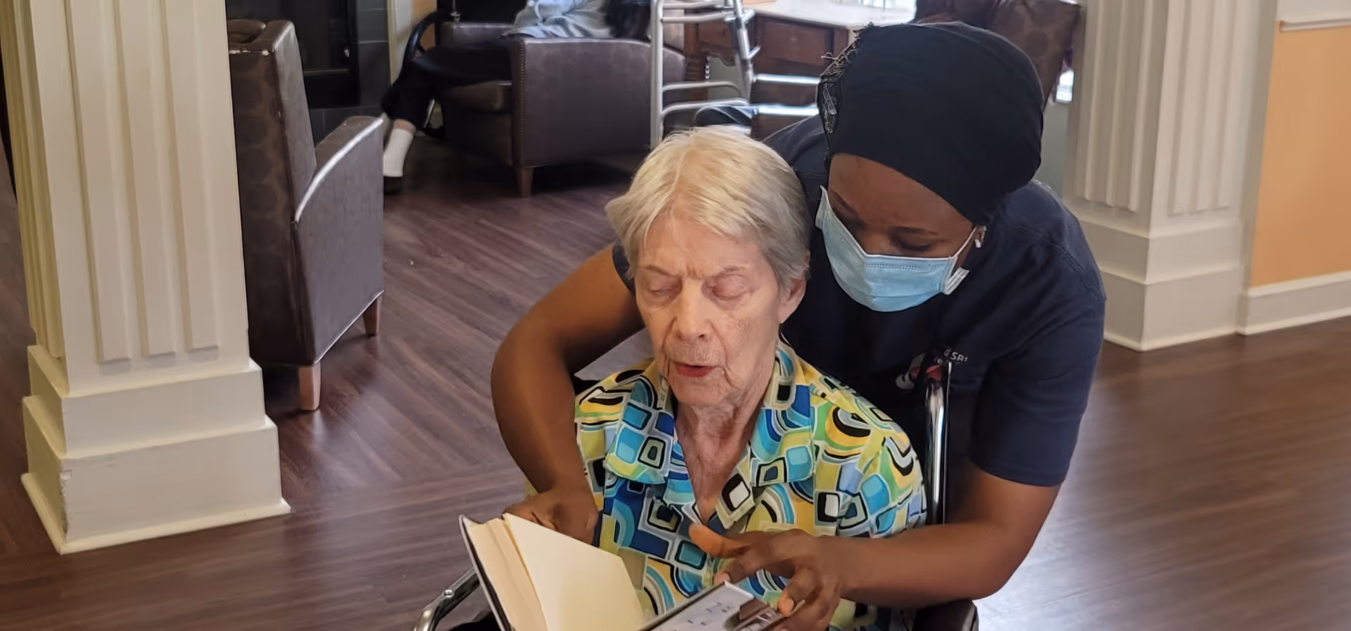 A masked caregiver leans over an elderly woman in a wheelchair to help her read a book in a senior living lounge.