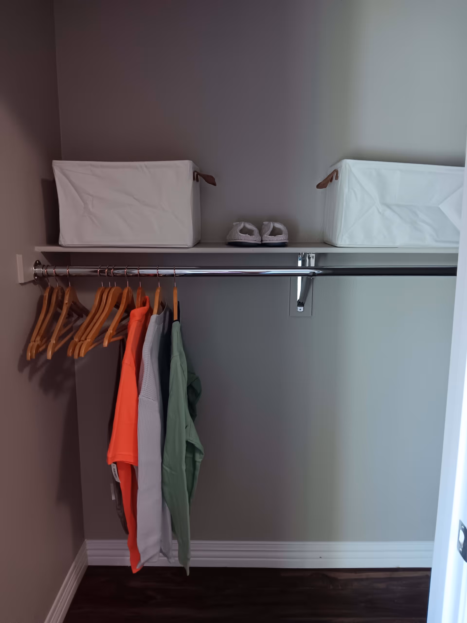 An open closet with a metal hanging rod holding several wooden hangers and a few pieces of clothing in various colors. Above the rod is a shelf with two white fabric storage bins and a pair of white slippers placed between them. The closet has light-colored walls and dark wood flooring.