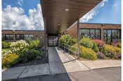 Entrance to a building with a covered walkway, surrounded by landscaped bushes and plants under a partly cloudy sky.