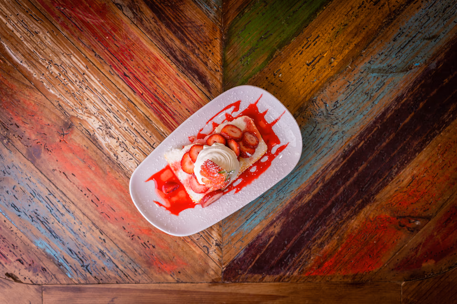 Top-down view of a strawberry-topped dessert with whipped cream and red sauce on a white plate on a colorful wooden table.