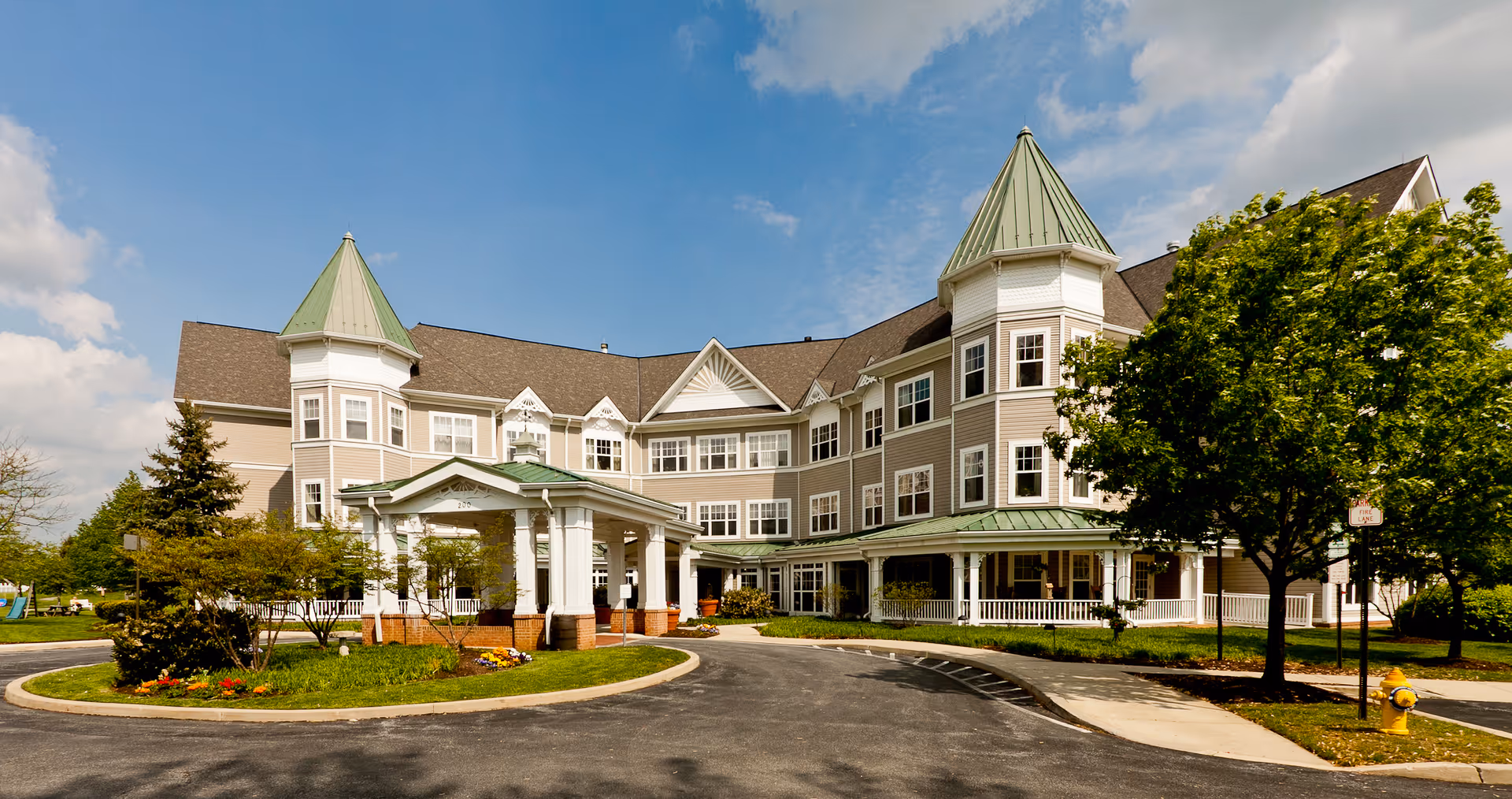 Exterior front view of a large senior living facility building with beige siding, green pointed roofs on two towers, multiple windows, a covered entrance with white columns, surrounded by trees and a circular driveway under a partly cloudy blue sky.