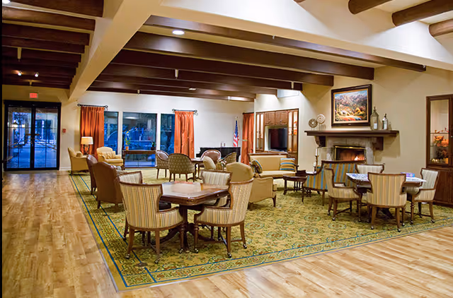 A spacious living room area in a senior living facility with wooden beams on the ceiling, a large patterned rug, multiple seating arrangements including armchairs and tables, a fireplace with a painting above it, and large windows with red curtains letting in natural light.