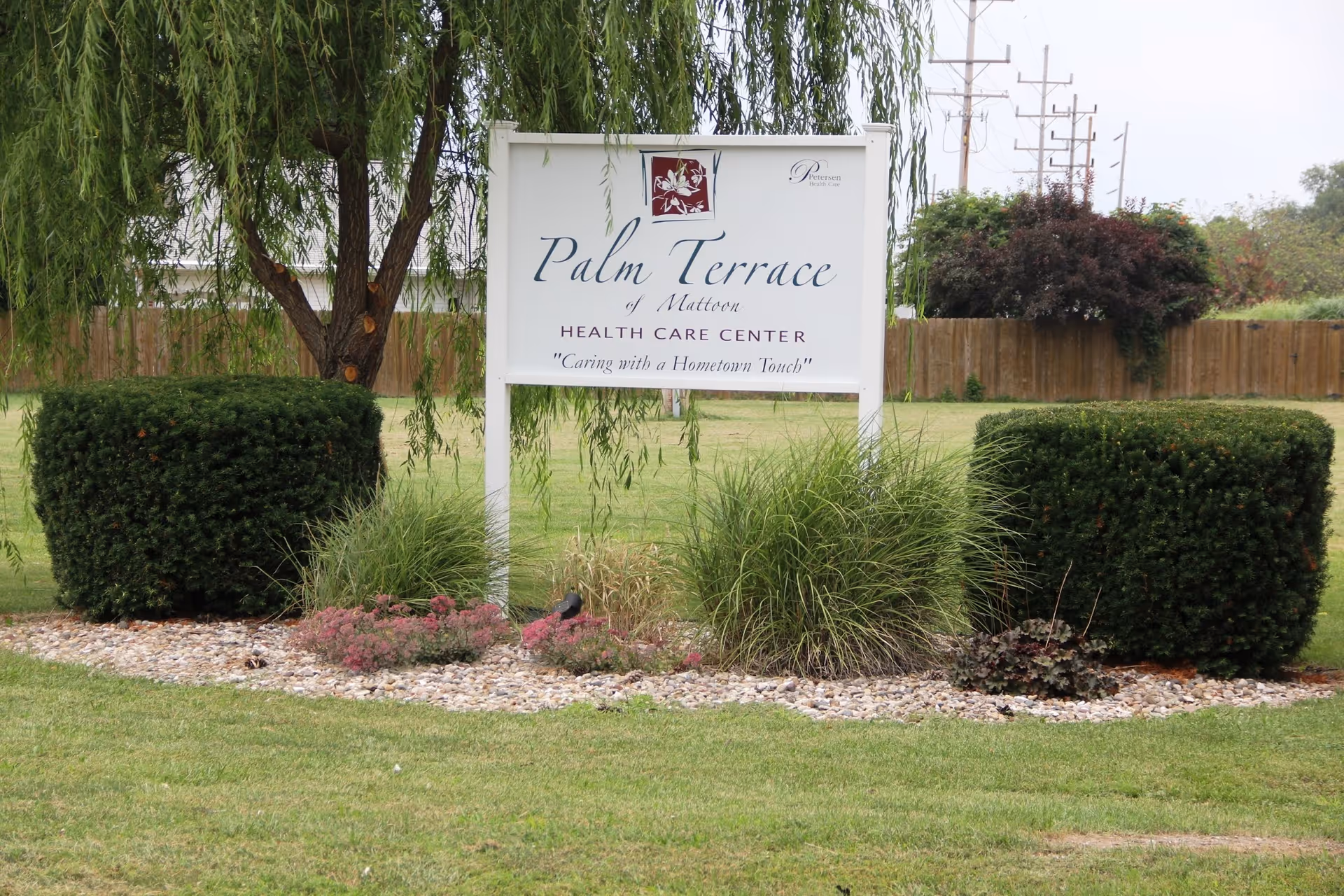 Outdoor view of a sign for Palm Terrace of Mattoon Health Care Center surrounded by neatly trimmed bushes, grass, and a tree with hanging branches.