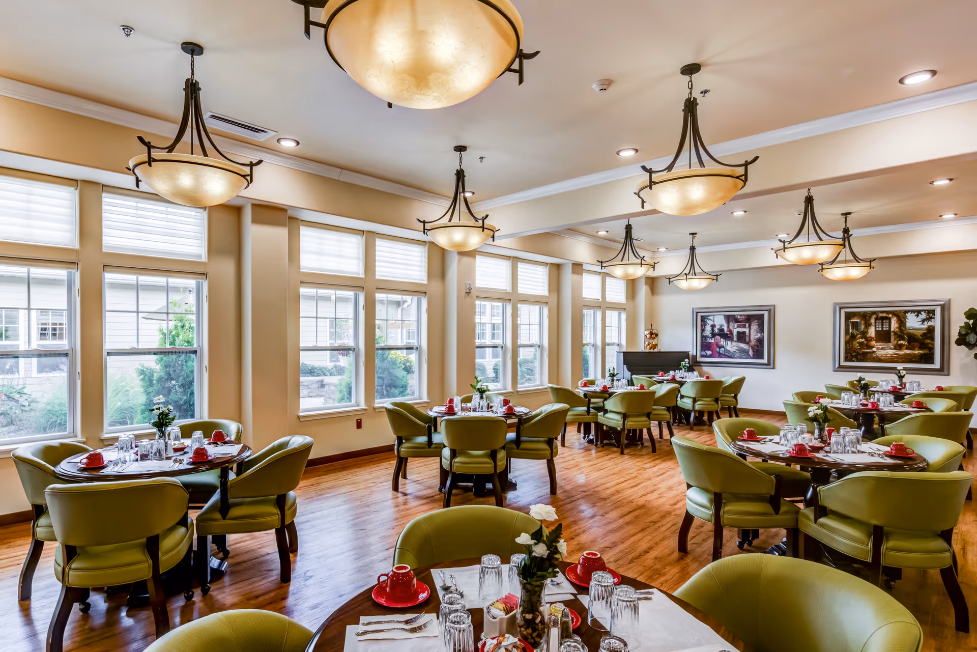 Dining room with round tables set for meals, green chairs, large windows and ornate hanging lights.