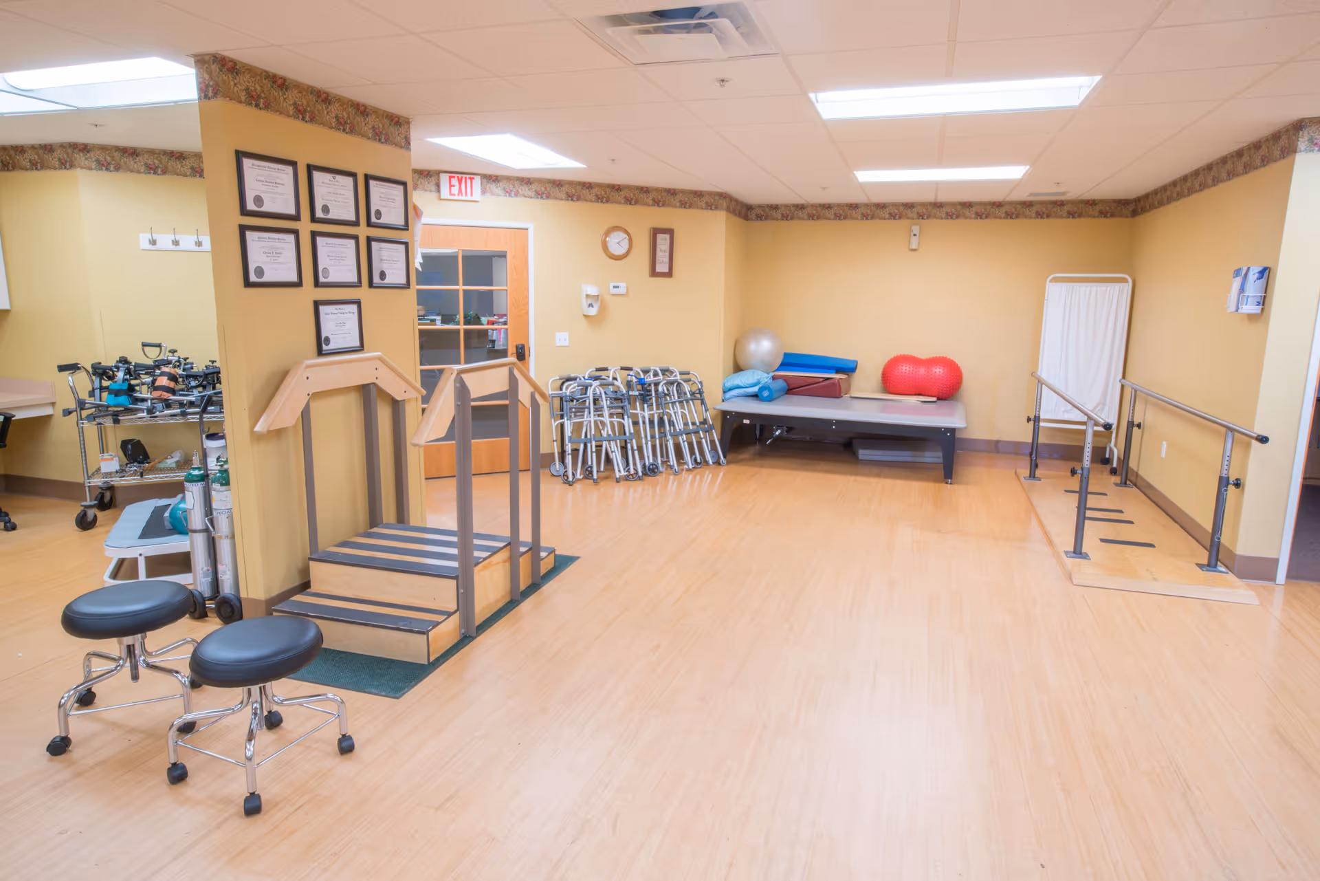 A physical therapy room with wooden flooring and beige walls. The room contains exercise equipment including parallel bars, a small set of stairs with handrails, walkers, exercise balls, foam rollers, and two black rolling stools. Several framed certificates are mounted on a wall near a door with a glass window. The room is well-lit with ceiling lights.