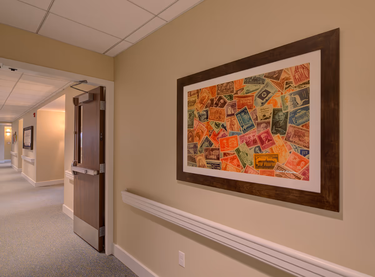 Interior hallway of The Lodge at Grand Junction with beige walls, blue patterned carpet, and a wooden door partially open. A large framed artwork featuring a collage of colorful vintage postage stamps is mounted on the wall.
