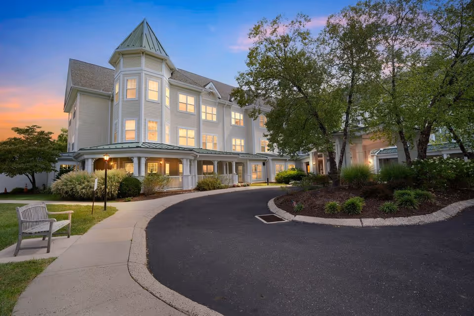 Exterior view of a multi-story senior living facility building at sunset with a curved driveway, landscaped garden area with trees and shrubs, a bench on the sidewalk, and warm lights glowing from the windows.