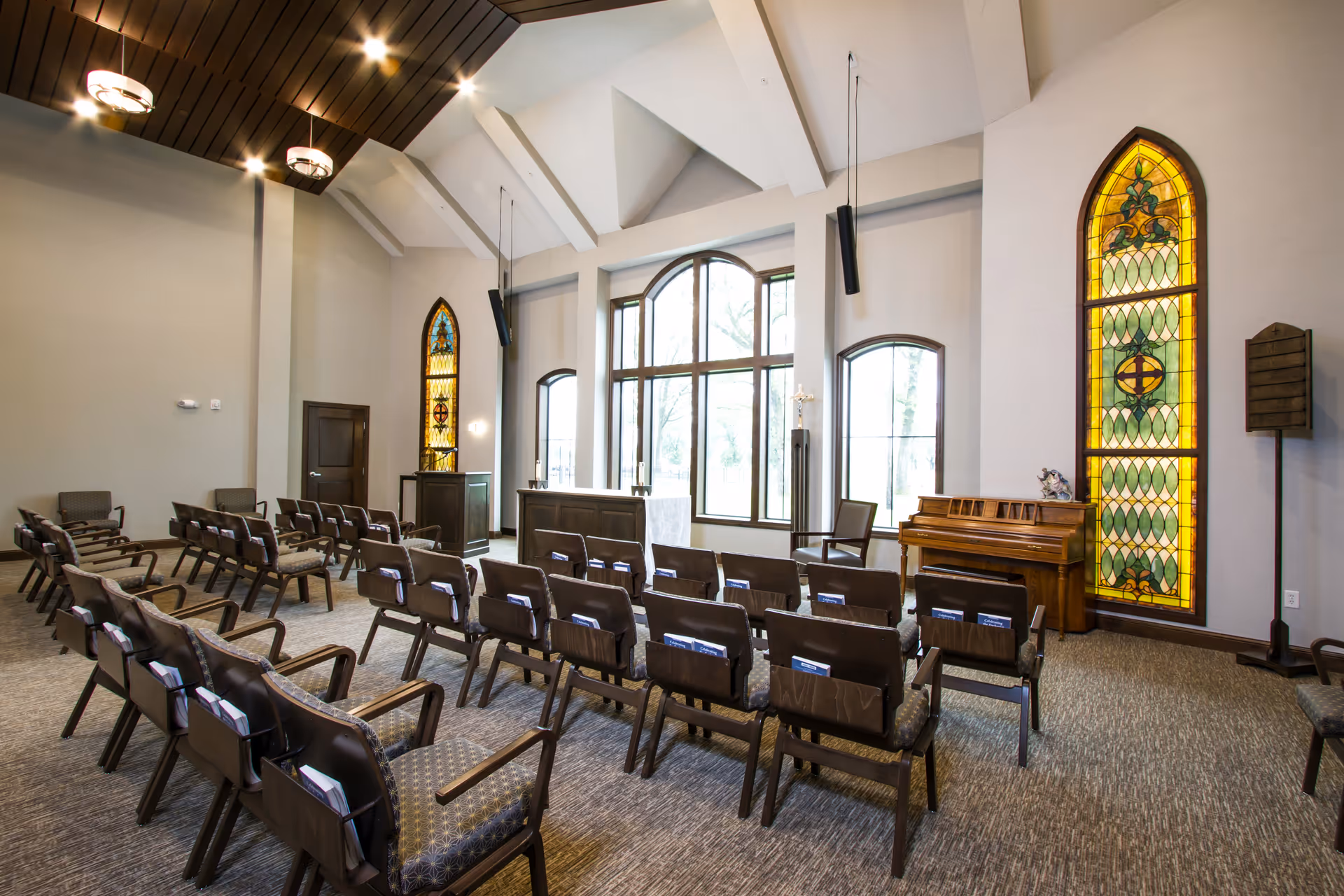 Interior view of a chapel or worship room with rows of chairs facing an altar. The room features large arched windows, two tall stained glass windows on either side, a wooden piano, and a high ceiling with modern light fixtures.