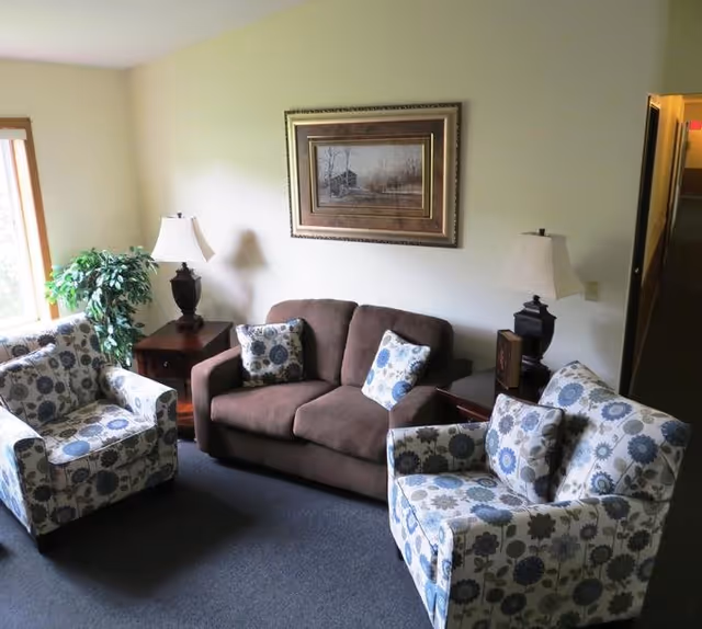 A cozy living room area with a brown loveseat and two floral-patterned armchairs arranged around two wooden side tables with matching lamps. A framed painting hangs on the light-colored wall above the loveseat, and a green potted plant is near a window letting in natural light.