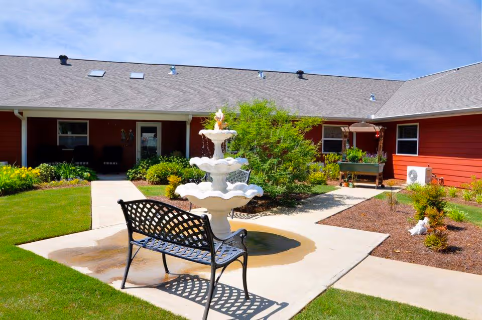 Outdoor courtyard area of a senior living facility with a three-tiered white fountain in the center, a black metal bench facing the fountain, green grass, landscaped plants, and a red building with a gray roof in the background under a clear blue sky.