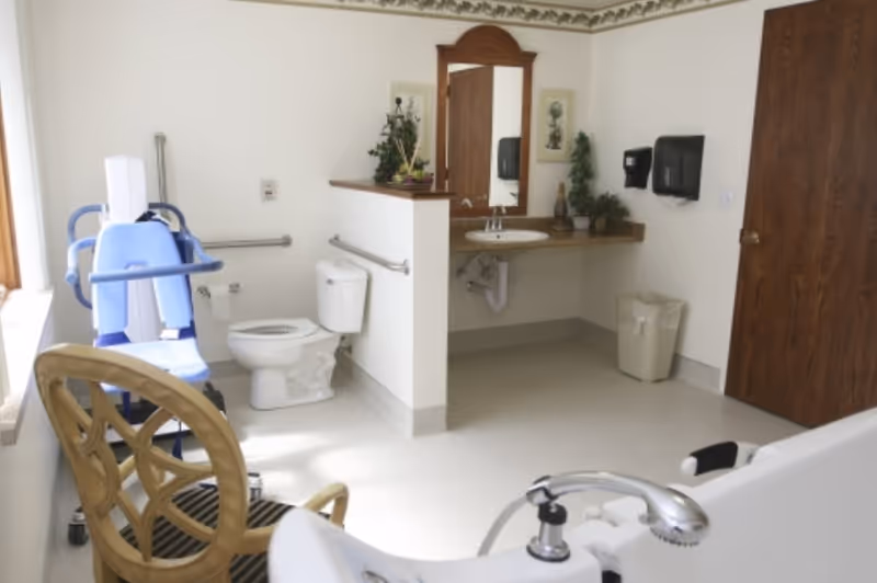 A spacious, well-lit bathroom in a senior living facility with a toilet, a sink with a large mirror above it, grab bars for accessibility, a wooden chair, a trash bin, and a blue patient lift chair near the window.