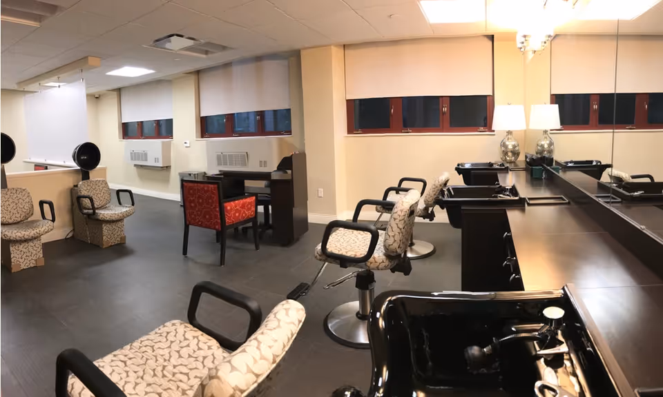Interior view of a hair salon area with several styling chairs, wash basins, a large mirror on the wall, and a desk with a red chair. The room has beige walls, windows with blinds, and overhead lighting.