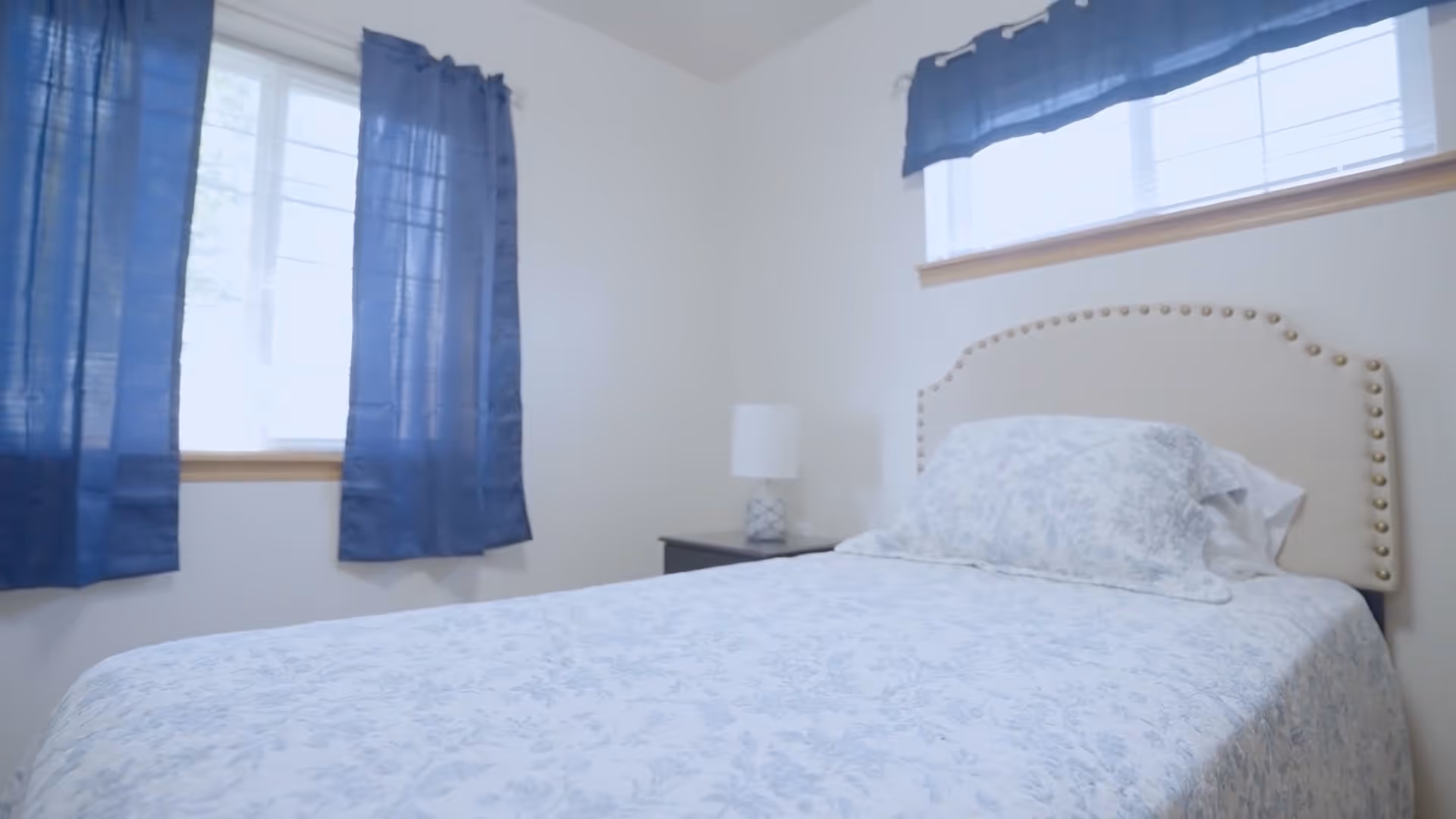 A bedroom with a bed covered in a light blue floral patterned bedspread and matching pillow. The bed has a beige upholstered headboard with nailhead trim. There is a small bedside table with a white lamp. The room has two windows with blue curtains and white walls.