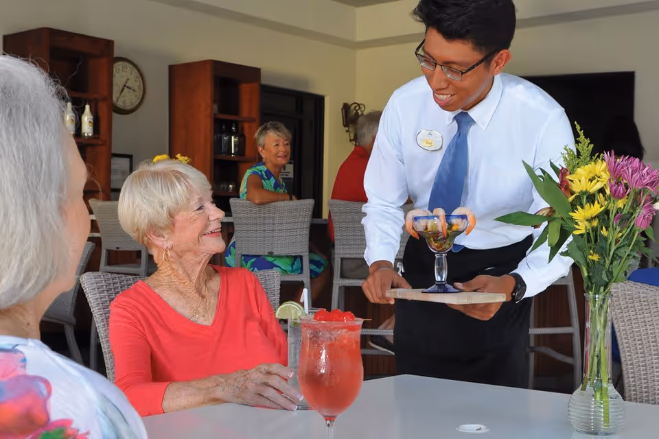 A smiling waiter serving a shrimp cocktail to an elderly woman in a red top seated at a table with a pink drink and a glass of water. Other elderly people are seated in the background in a dining area with wooden shelves and a clock on the wall. A vase with colorful flowers is on the table.