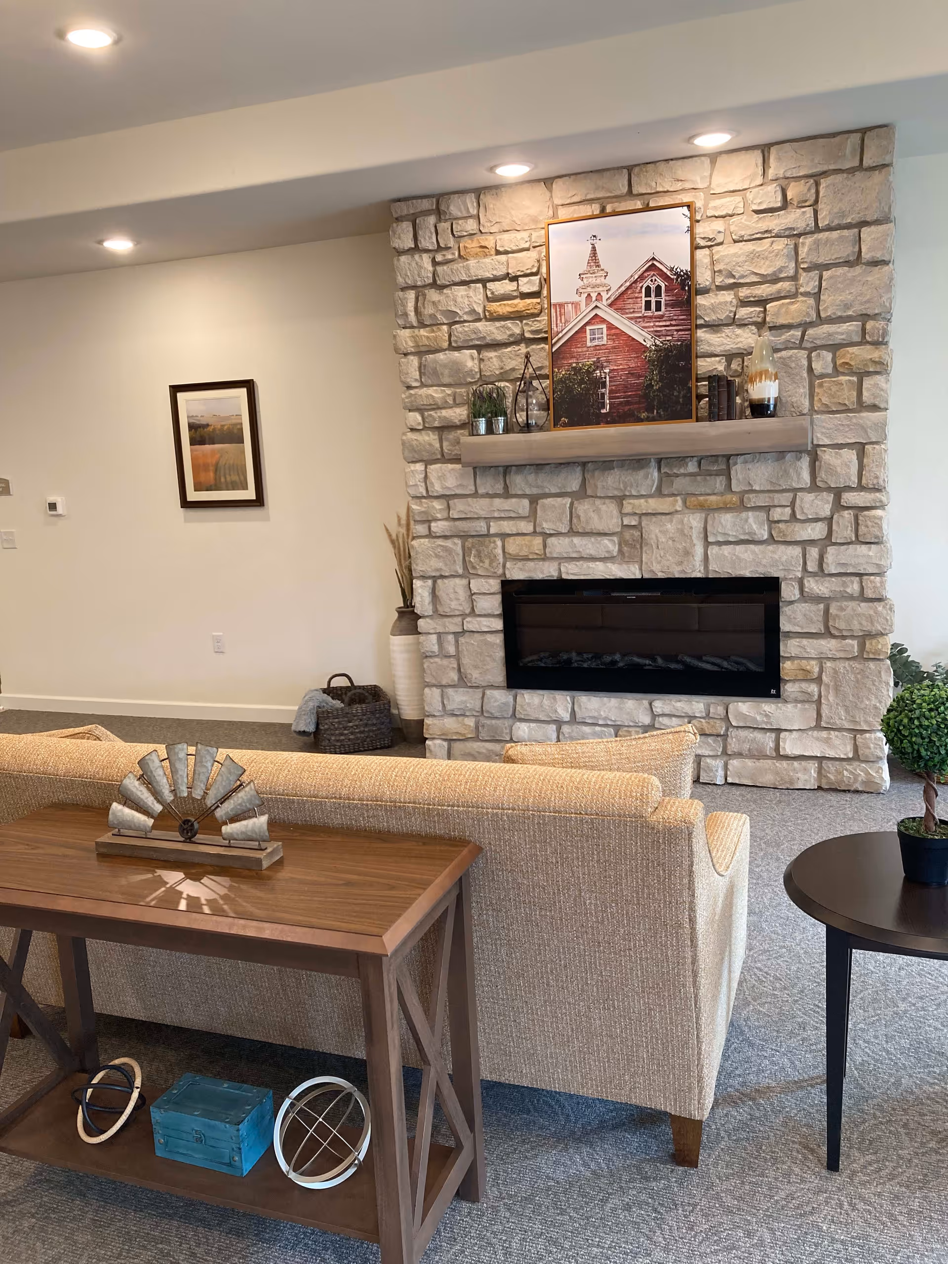 A cozy living room area featuring a beige sofa facing a stone fireplace with a wooden mantel. On the mantel, there is a framed picture of a red building with a steeple, along with decorative items including plants and books. In front of the sofa is a wooden console table with decorative objects, and to the right is a round side table with a small potted plant. The room has recessed ceiling lights and neutral-colored walls and carpet.