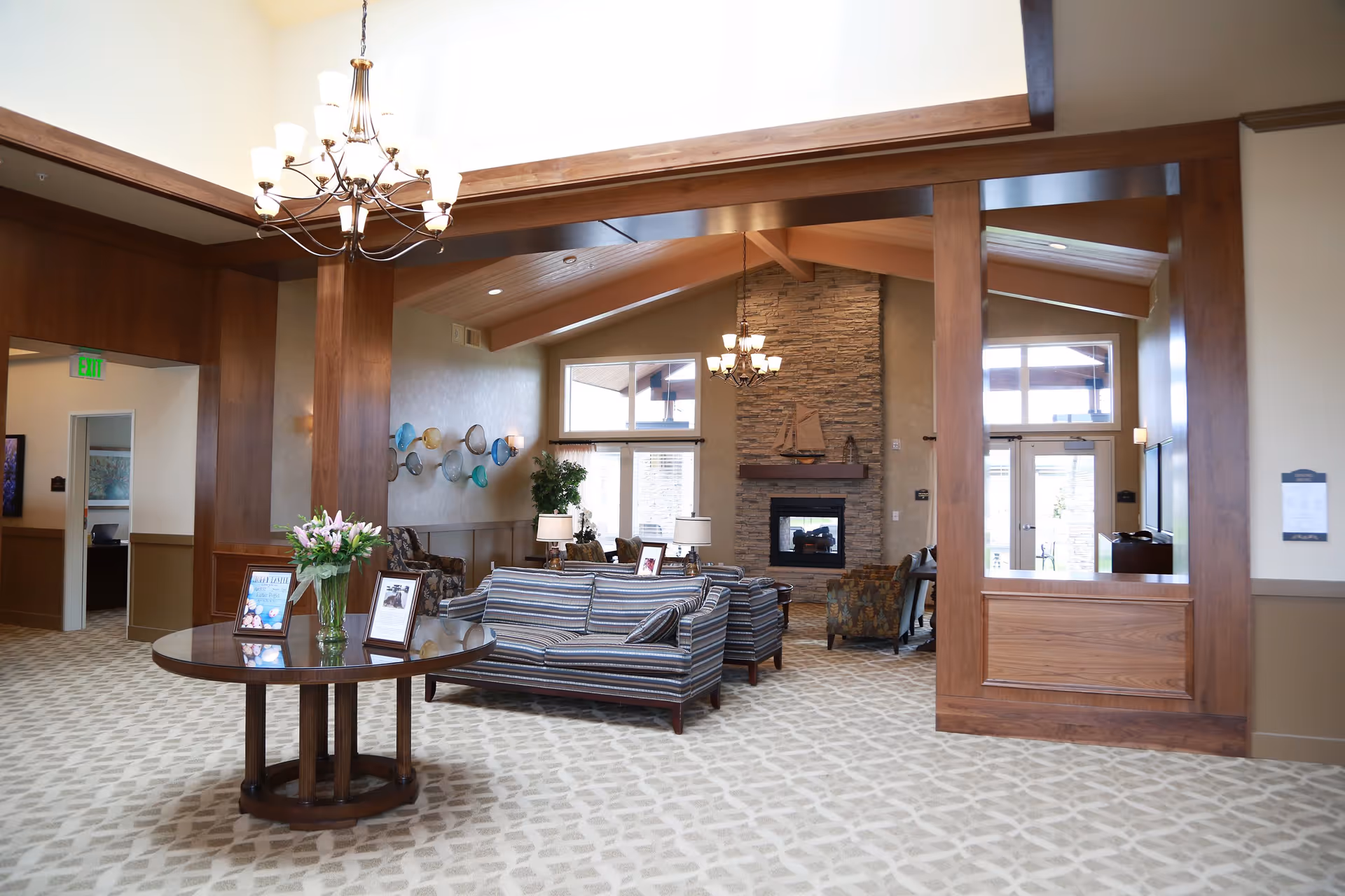 A spacious and well-lit living room area in Silverado Bellingham Memory Care Community featuring a high vaulted ceiling with wooden beams, a stone fireplace with a decorative sailboat on the mantel, striped sofas, patterned armchairs, a round wooden table with a vase of flowers and framed pictures, and wall art with colorful glass bowls. Large windows and glass doors allow natural light to fill the room.