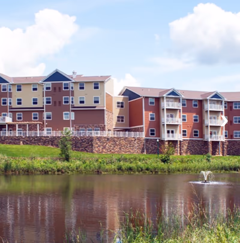 Exterior view of a multi-story senior living facility building with beige, brown, and red siding, balconies, and a stone foundation. In the foreground, there is a pond with a small fountain and green grass surrounding it under a partly cloudy sky.
