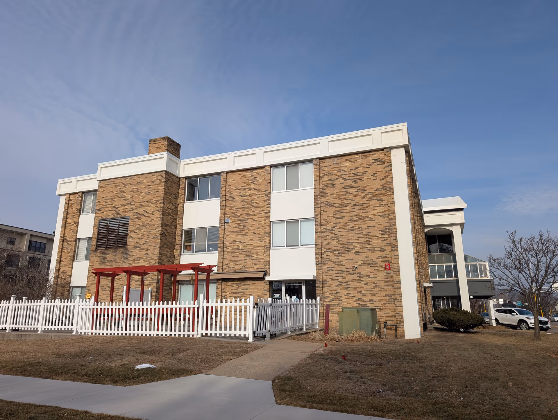 Exterior view of a three-story brick and white panel building with a white picket fence and a red pergola in front. There is a sidewalk leading to the entrance, some parked cars on the right, and a clear blue sky above.