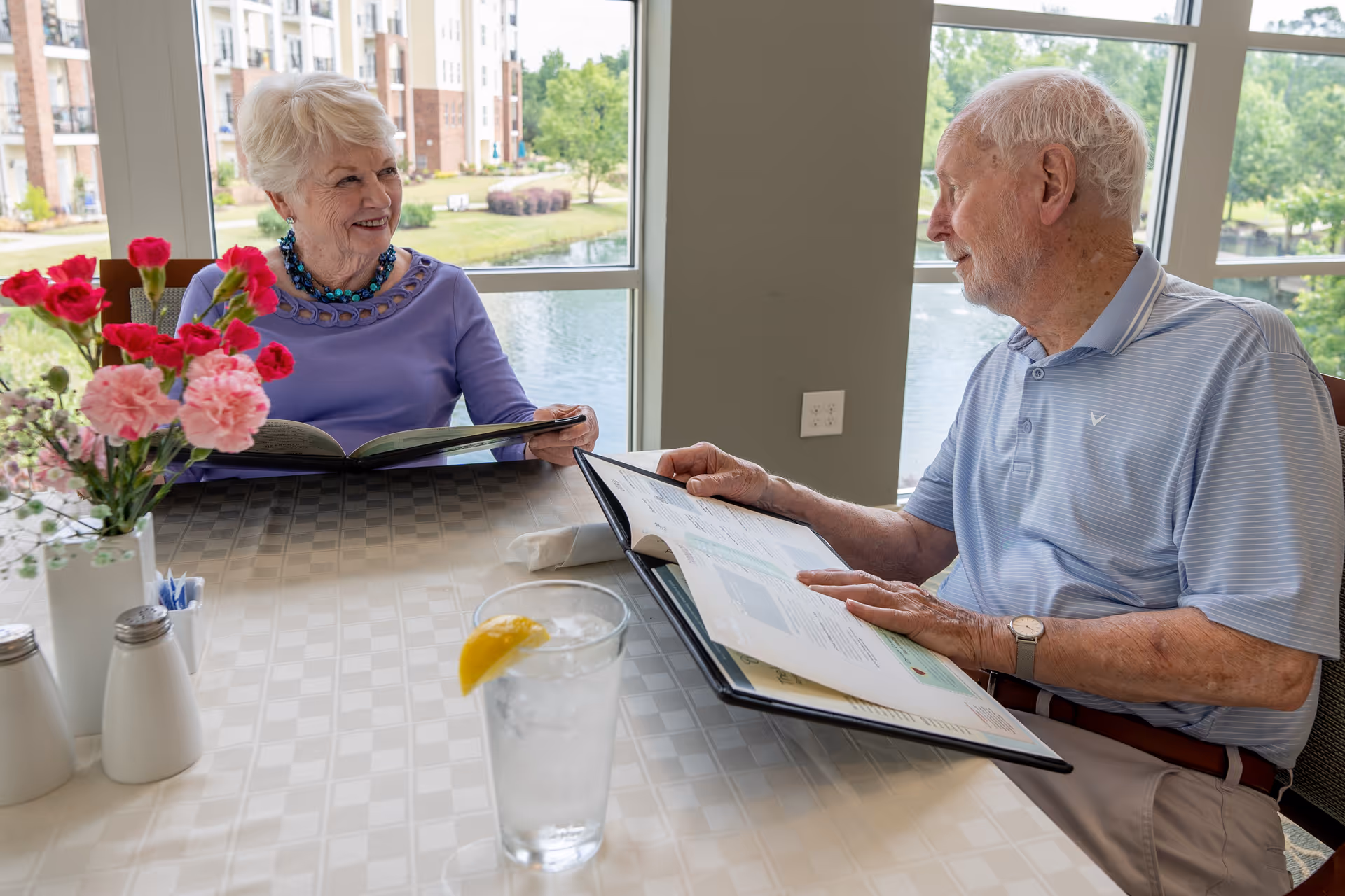 An elderly man and woman sit at a table in a dining area with large windows showing a pond and greenery outside. Both are looking at menus and smiling at each other. On the table are a glass of water with a lemon slice, a vase with pink and red flowers, and salt and pepper shakers.