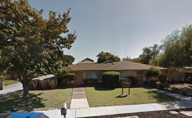 Single-story residential building with a brown roof and beige walls, surrounded by a green lawn and trees. A concrete walkway leads from the sidewalk to the front entrance. A sign is placed on the lawn near the entrance. A blue car is partially visible in the lower left corner.