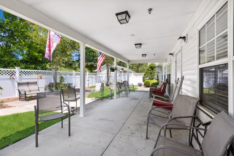 Covered outdoor patio area with multiple chairs arranged along the wall and open space, American and decorative flags hanging from the ceiling, white fence surrounding a green lawn with trees and shrubs in the background.