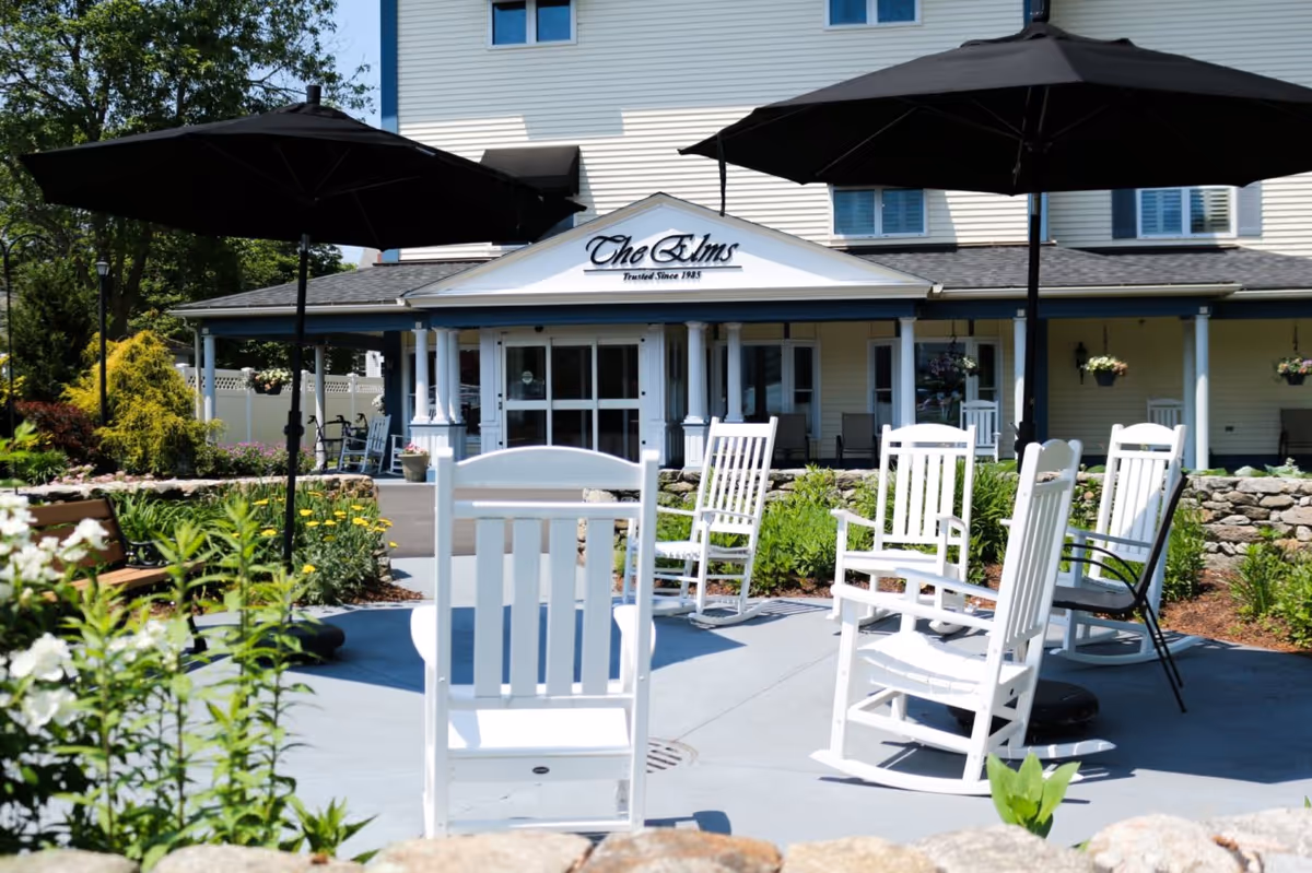 Outdoor seating area with white rocking chairs and black umbrellas in front of a building with a sign that reads 'The Elms'. The area is surrounded by greenery and flowers with a stone wall in the foreground.