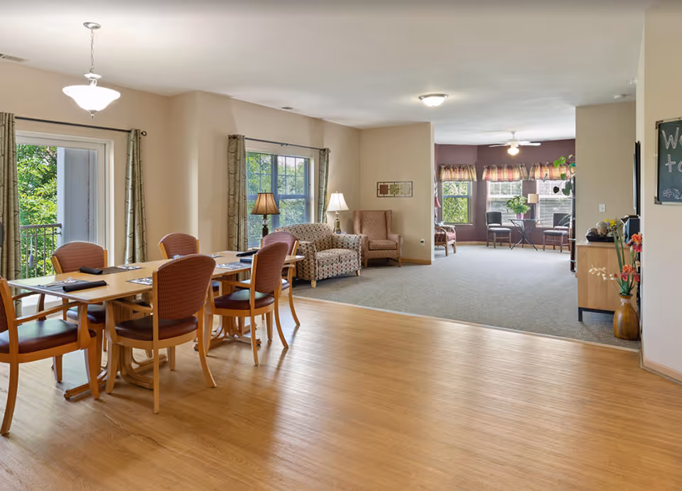 A spacious and well-lit common area in a senior living facility featuring a dining table with six chairs set with placemats and napkins on the left side, a cozy sitting area with patterned armchairs and lamps near windows in the center, and another seating area with chairs and a small table near large windows in the background. The room has wood flooring in the dining area and carpet in the sitting areas, with light-colored walls and large windows letting in natural light.
