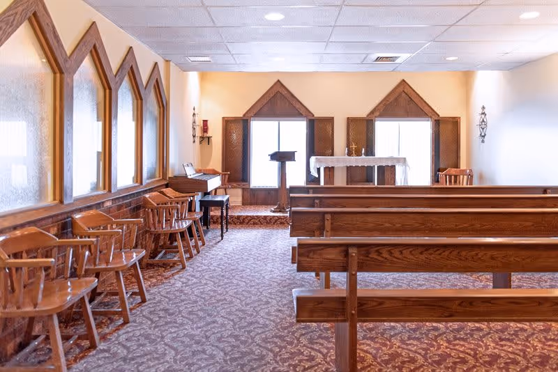 Interior view of a small chapel or prayer room with wooden pews and chairs arranged facing an altar with a lectern and a table covered with a white cloth. The room has patterned carpet flooring, beige walls, and windows with wooden frames.