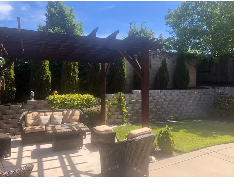 Sunlit backyard patio with a wooden pergola, wicker seating and cushions, a small lawn, and a stone retaining wall with trees.
