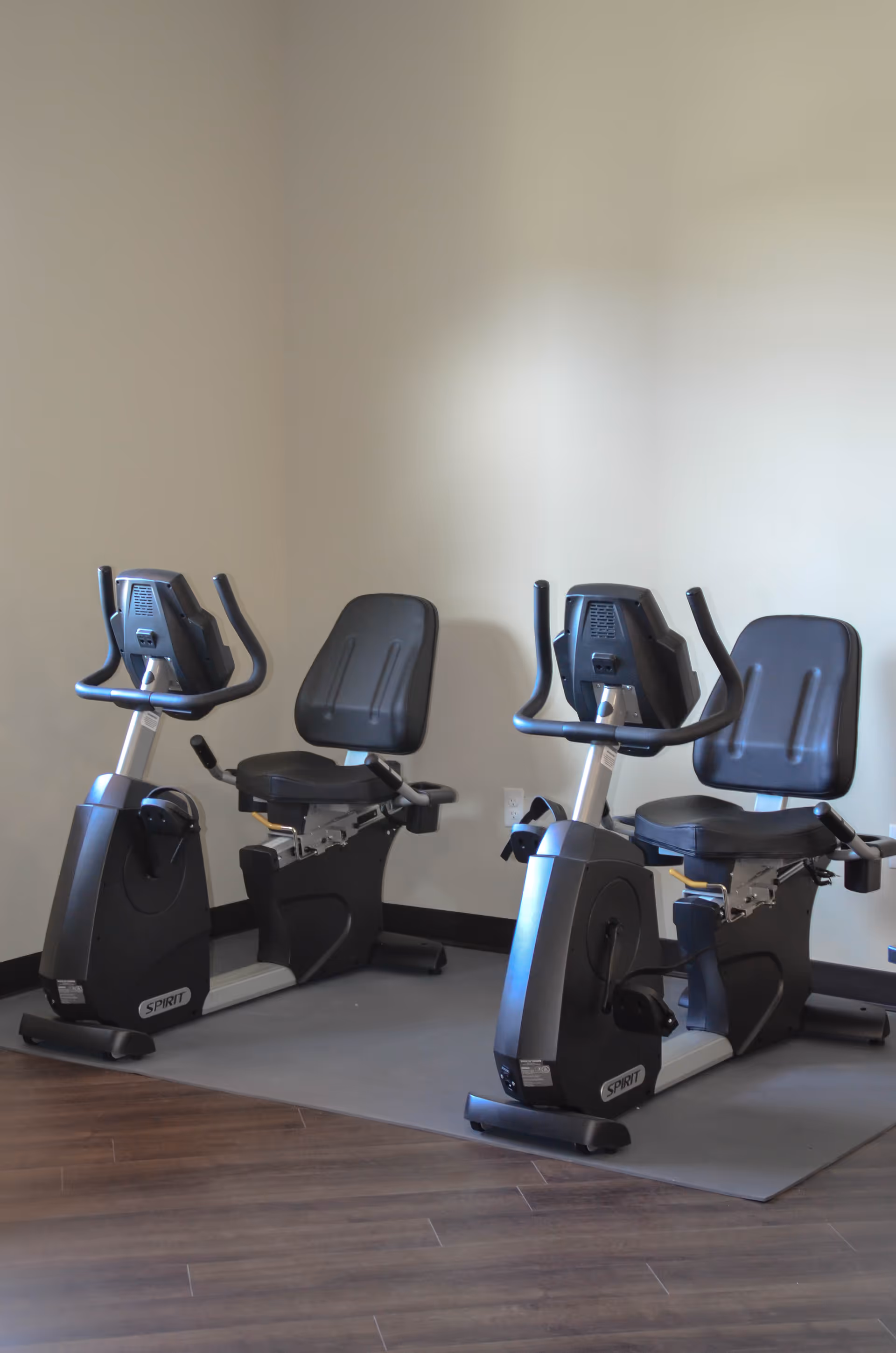 Two stationary recumbent exercise bikes positioned side by side on a mat in a room with wooden flooring and plain light-colored walls.