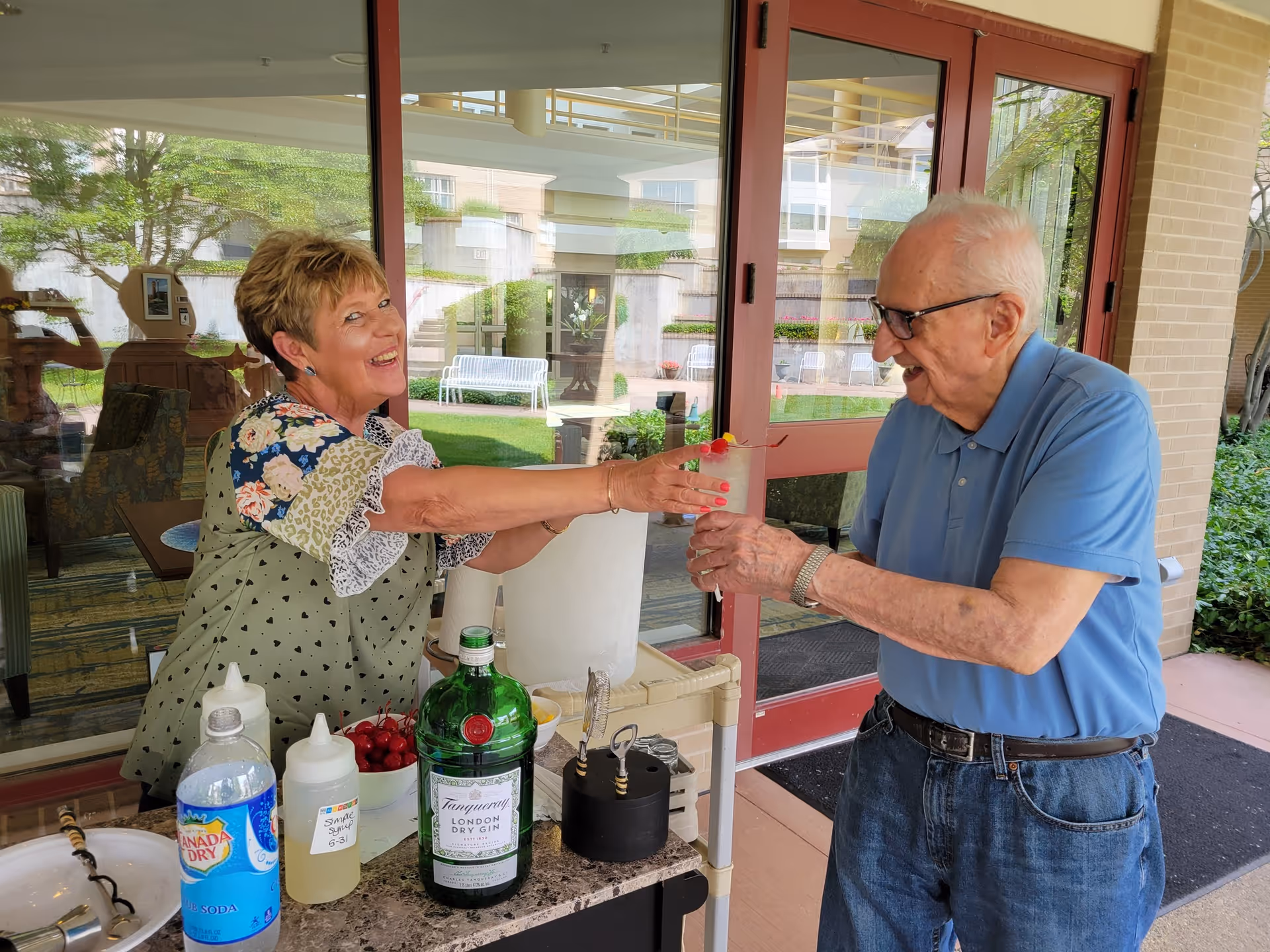 An elderly man in a blue polo shirt and jeans is receiving a drink from a smiling woman standing behind a small outdoor bar setup. The woman is wearing a green patterned blouse with floral and lace details. On the bar counter, there are bottles including a large bottle of Tanqueray London Dry Gin, a bottle of Canada Dry club soda, a bowl of cherries, and other drink-making supplies. They are outside near a building with large glass doors and windows, with greenery and benches visible in the background.