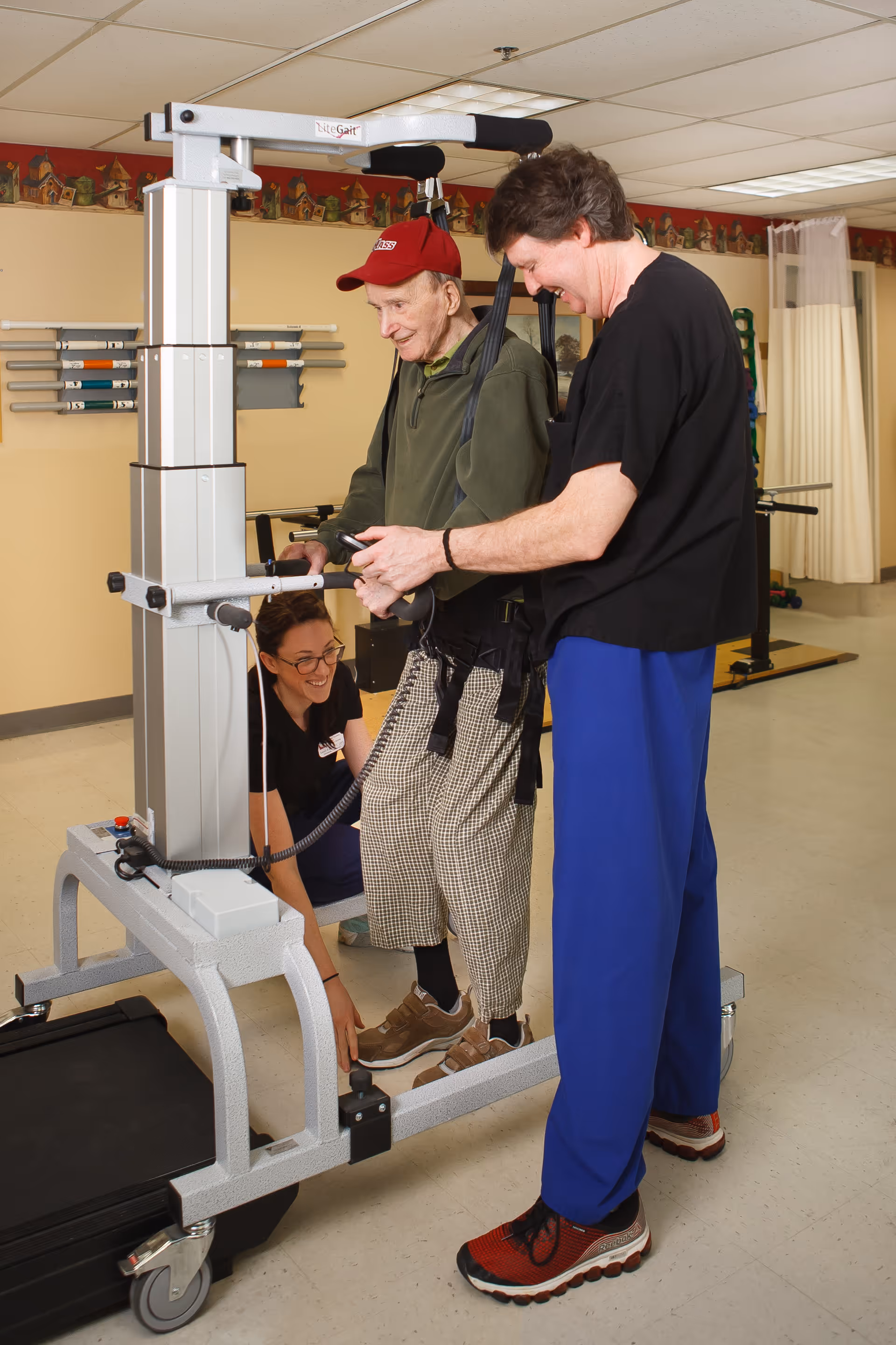 An elderly man wearing a red cap and checkered pants is supported by a standing lift device while two caregivers assist him in a rehabilitation or therapy room with exercise equipment in the background.