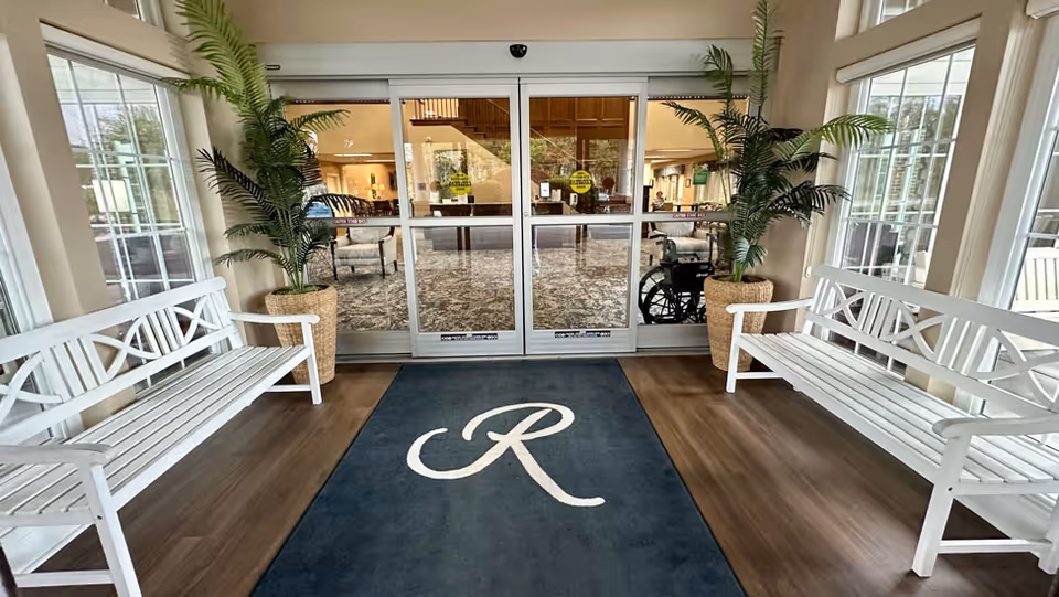 Entryway of a senior living facility with two white benches on either side, large potted plants, a dark blue rug with a white letter 'R', and glass sliding doors leading inside where a wheelchair and seating area are visible.