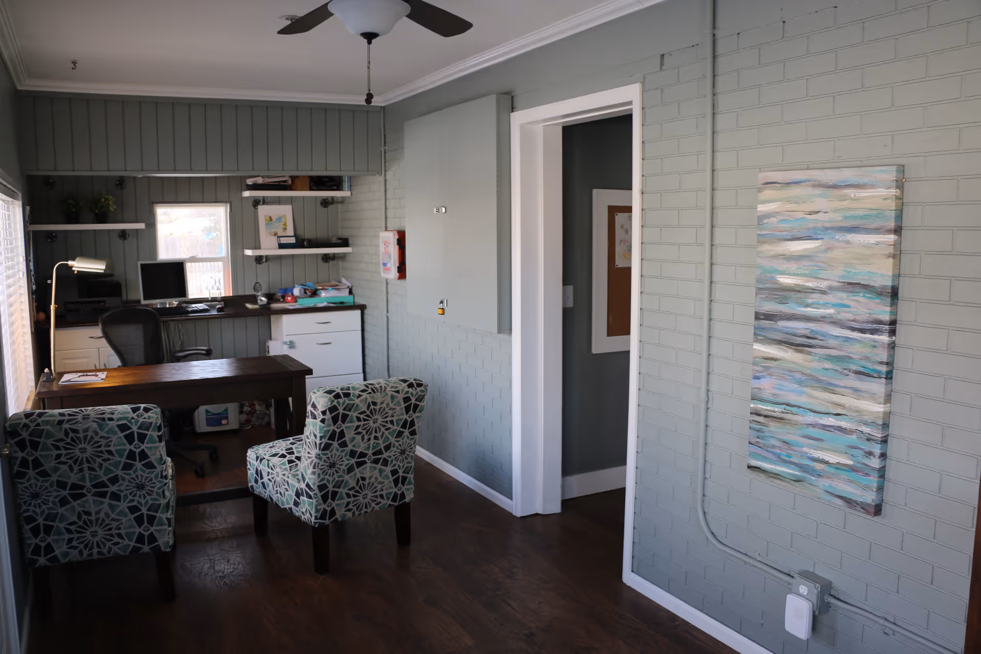 An office room with a wooden desk, two patterned upholstered chairs, a black office chair, a computer monitor, shelves with books and decor, a ceiling fan, and a large abstract painting on a gray brick wall.